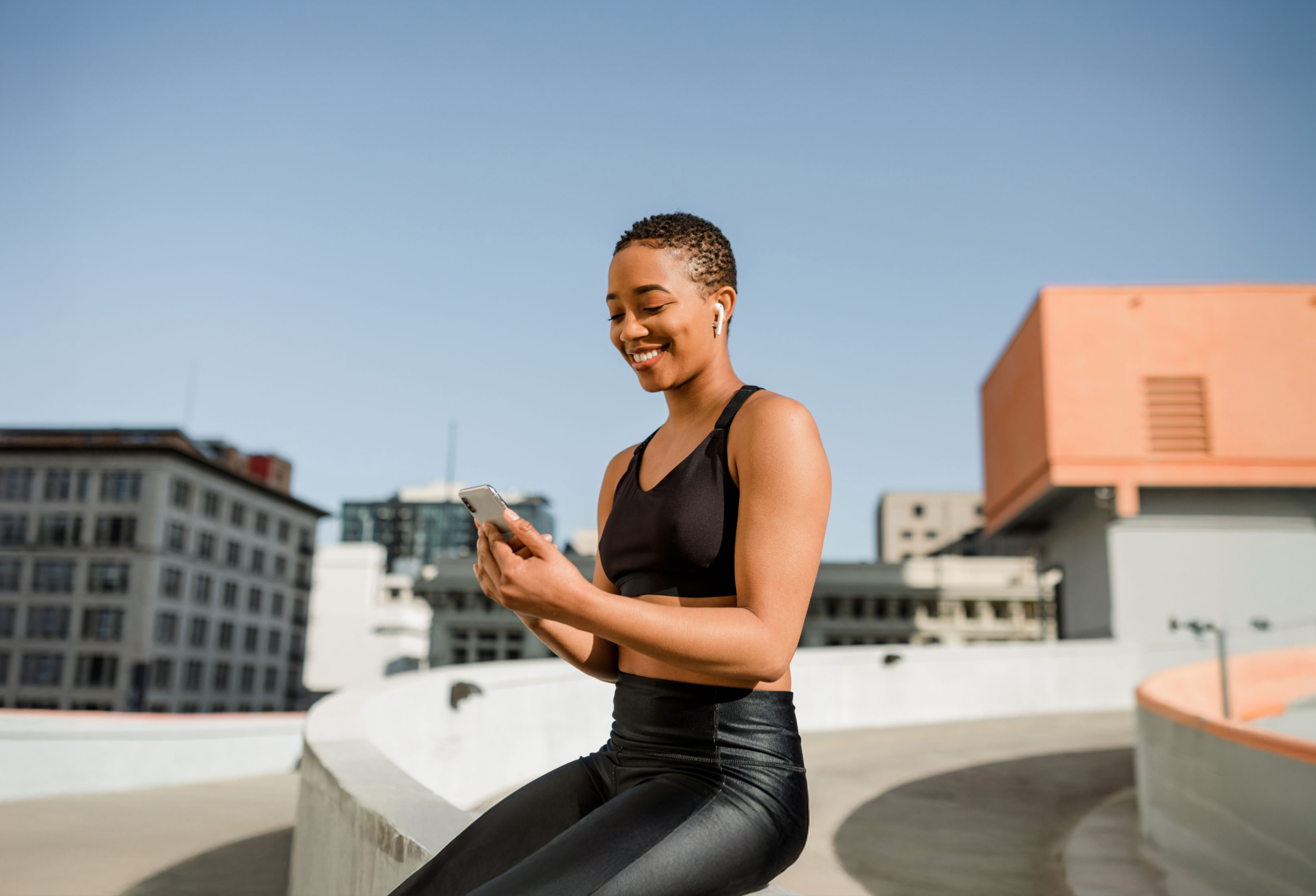 A young woman wearing black athletic wear is sitting outdoors on a rooftop in an urban setting. She is smiling while looking at her phone and wearing wireless earbuds. The background features city buildings and a clear blue sky.
